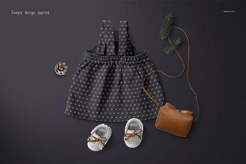 Front view of a baby dress mockup with a dark patterned fabric, displayed with baby shoes, a brown purse, pine sprigs, and a pinecone.