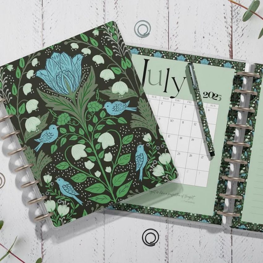 Top view of two spiral planners on a white wooden surface, one closed with a floral and bird pattern, the other open to a July calendar page.