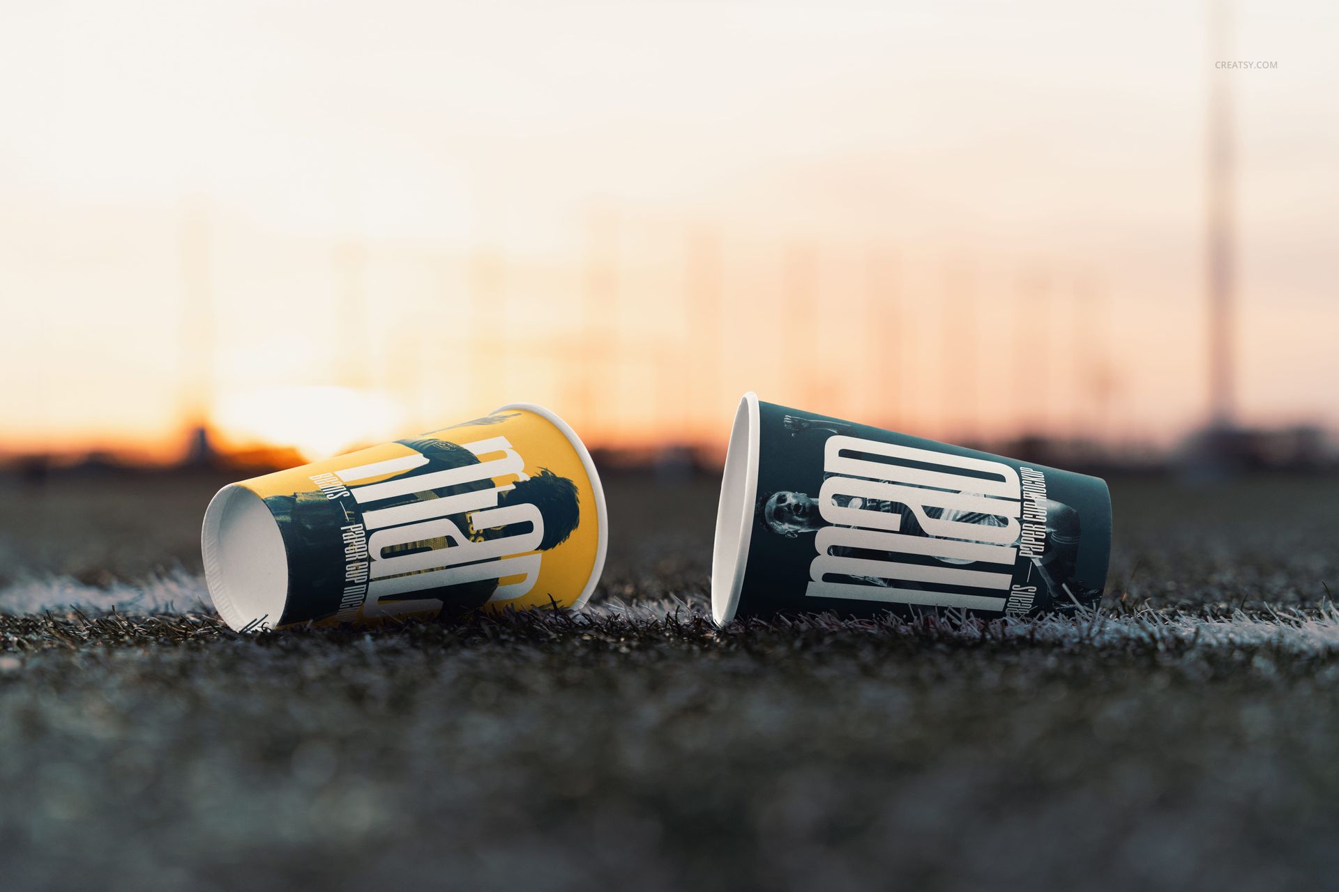 PSD mockup of drinkware showing two decorated paper cups on a stadium turf with a blurred sunset background.