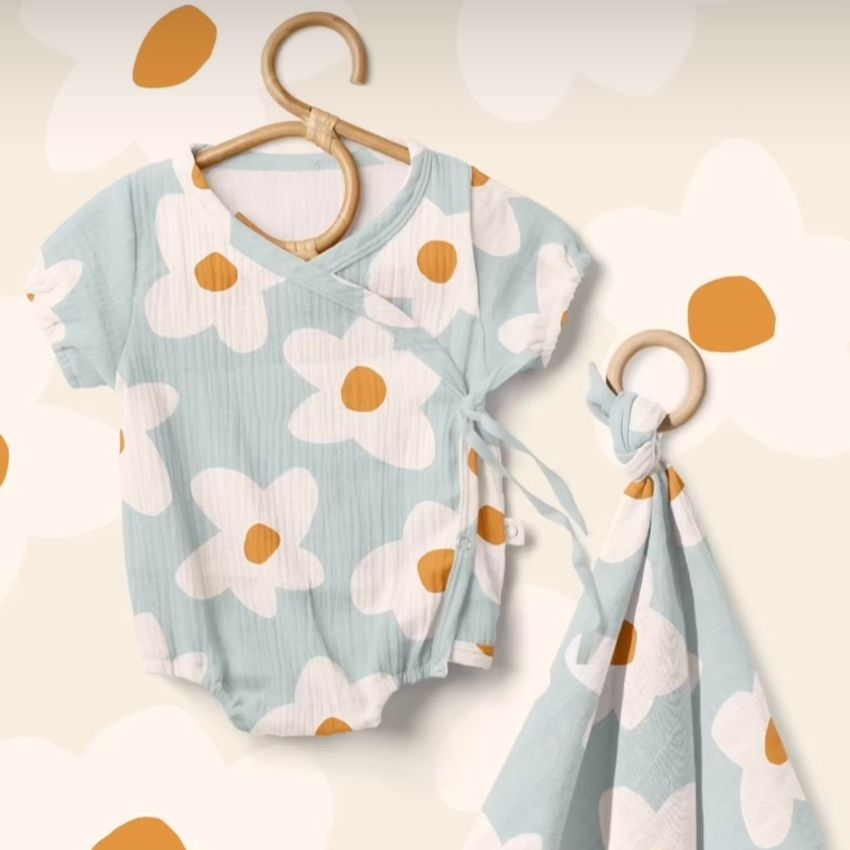 Muslin onesie mockup showing a short-sleeve baby bodysuit with a blue background and large white and orange floral pattern, displayed on a wooden hanger next to a matching knotted muslin cloth.