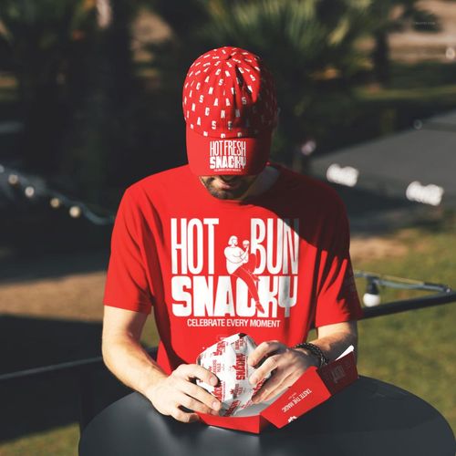 Man in red fast food themed t-shirt and cap sitting outdoors, holding a branded burger wrapper and box on a round table.