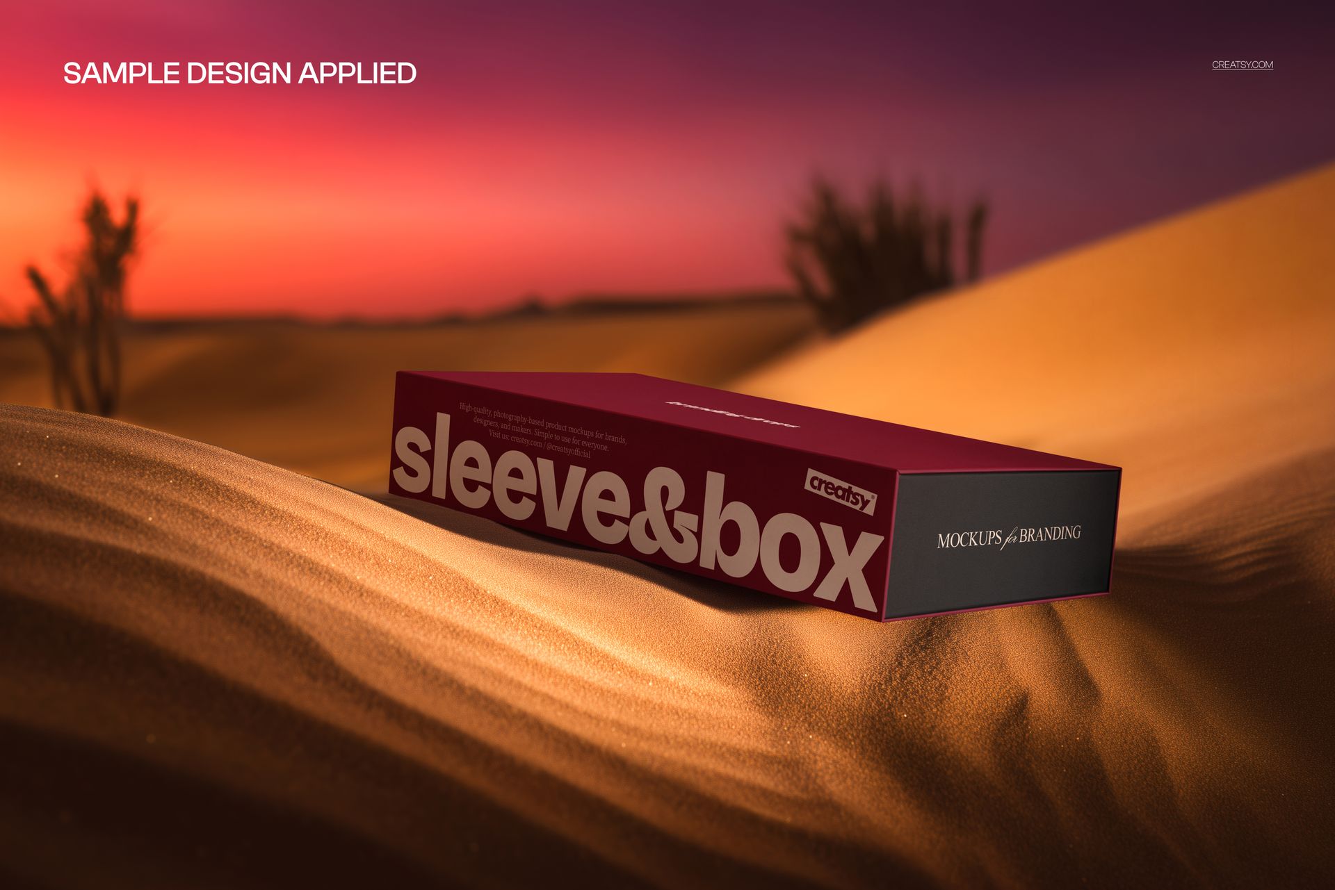 Mockup of a maroon rectangular packaging box with sleeve, resting on textured sand dunes under warm evening light.