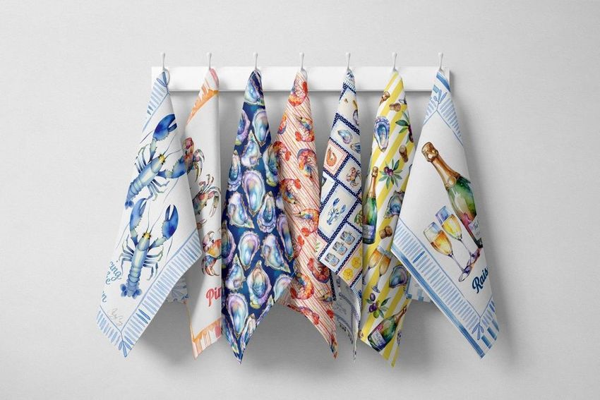 Hanging tea towels mockup showing seven colorful towels with coastal-themed illustrations, displayed on a white rack against a light gray wall.