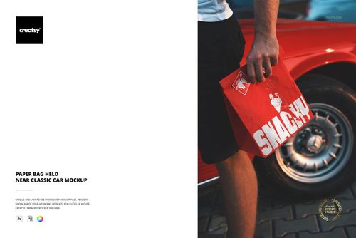 Hand holding a vibrant red paper bag near the wheel of a vintage red car on a paved surface, PSD mockup scene.