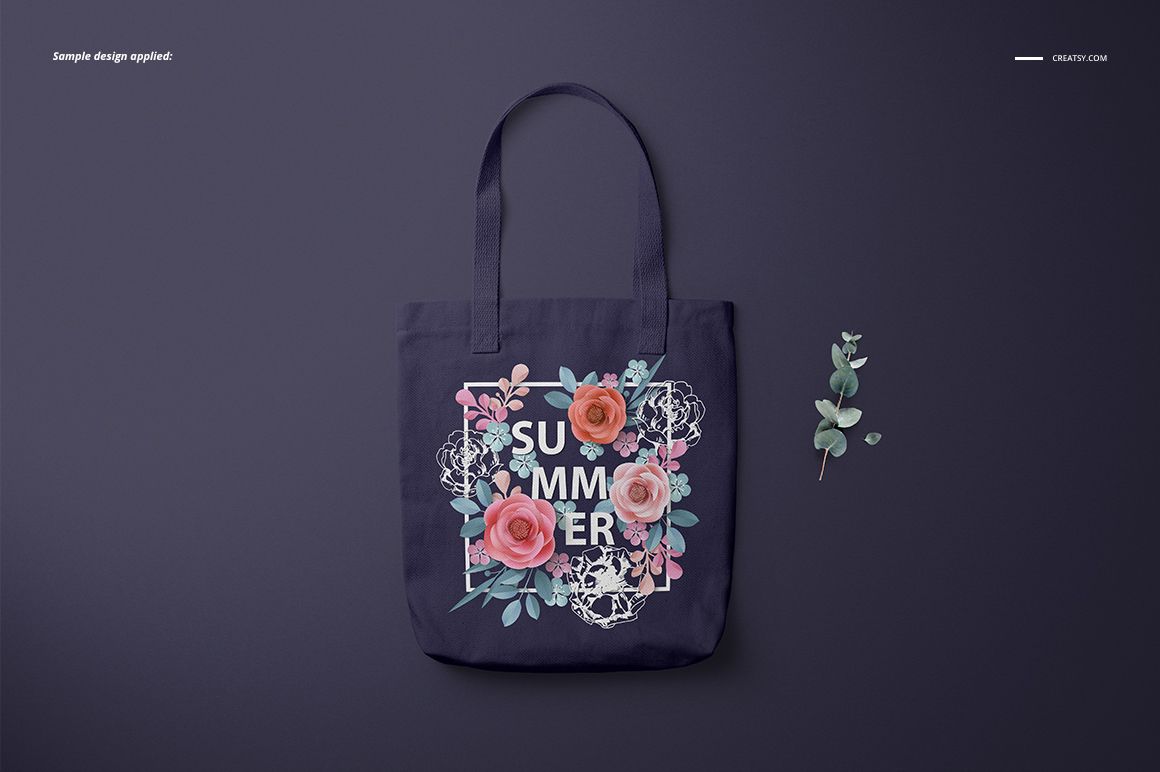 Front view of a polyester tote bag mockup with floral graphics and text, displayed on a dark surface with a sprig of greenery.