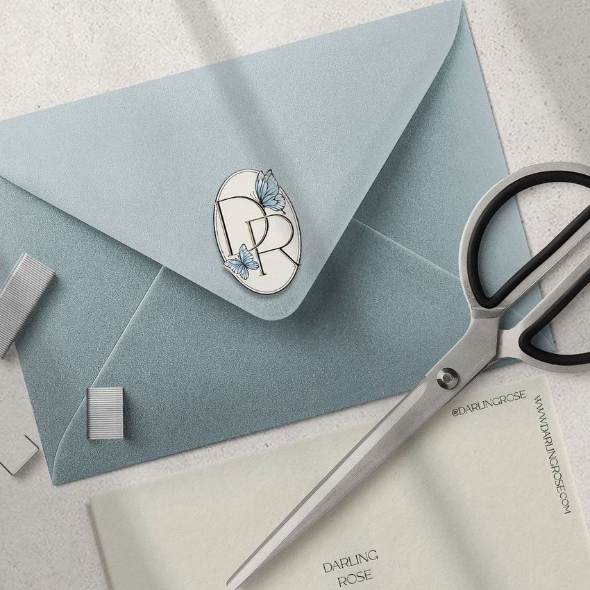 Folded card mockup showing a light blue envelope sealed with a monogram sticker and butterfly design, placed next to scissors and stationery.