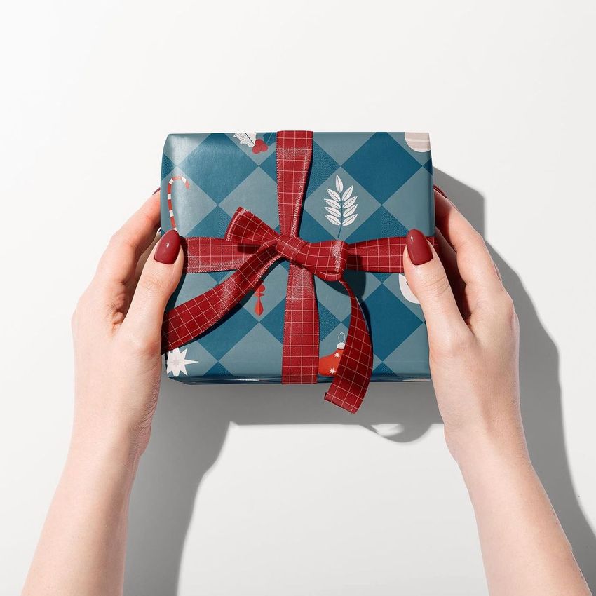 Gift box mockup wrapped in blue patterned paper with a red plaid ribbon, held by two hands against a white background.