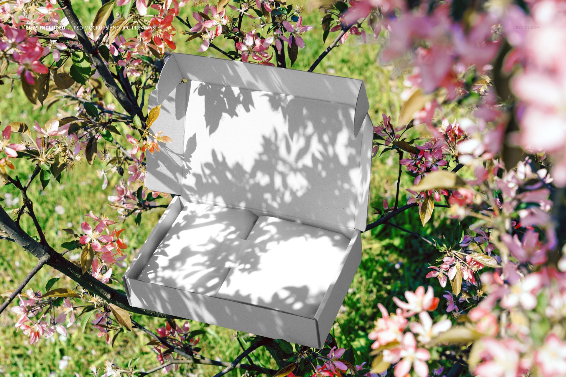 Rectangular mailing box mockup with open lid, placed among blooming spring branches with green grass visible.