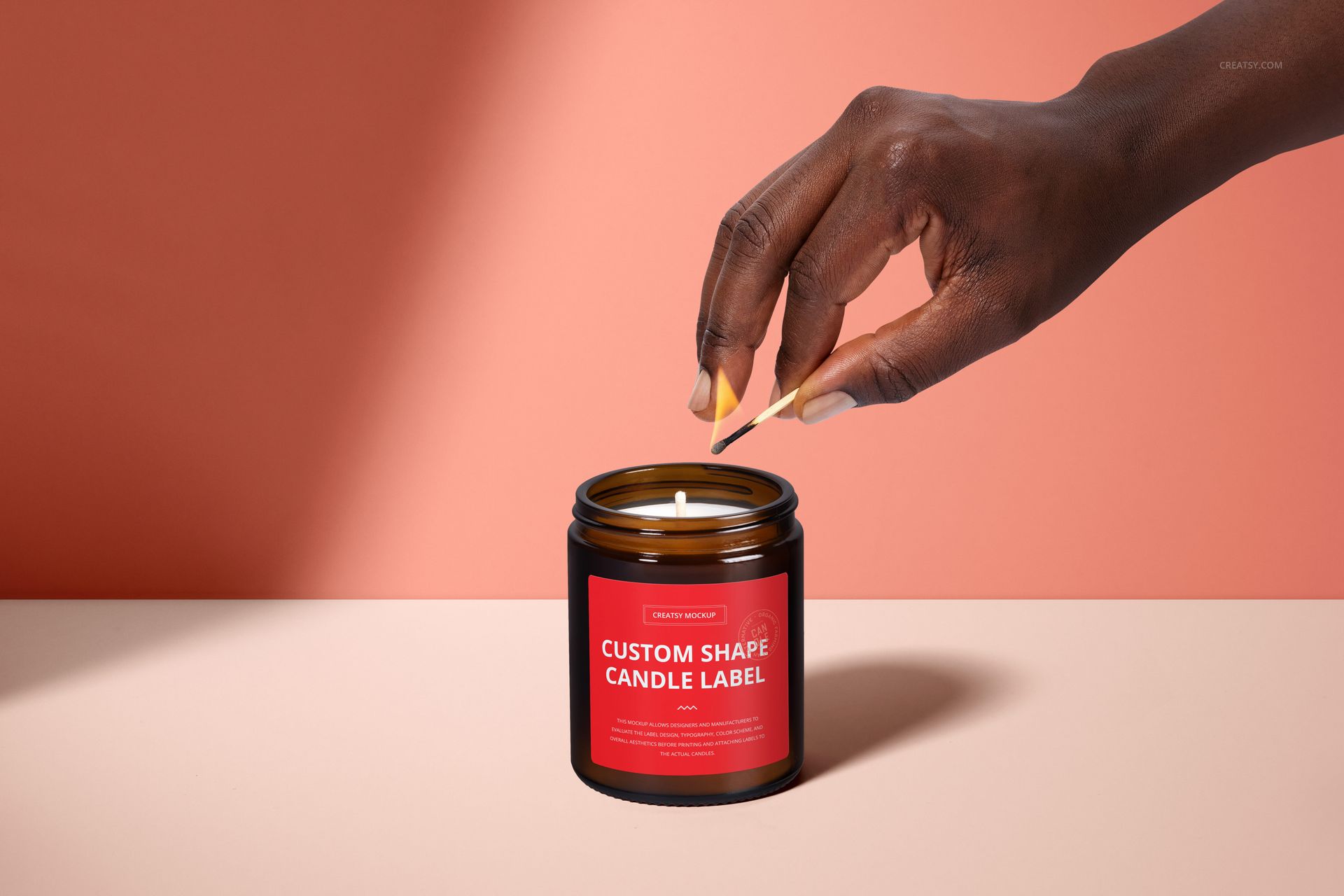 PSD mockup of a brown glass candle jar with red label, cotton wick, and hand igniting the candle on a neutral surface.