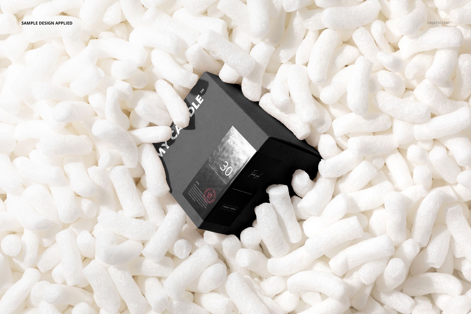 Square box with dark design partially submerged in white packaging peanuts, viewed from above in a mockup scene.