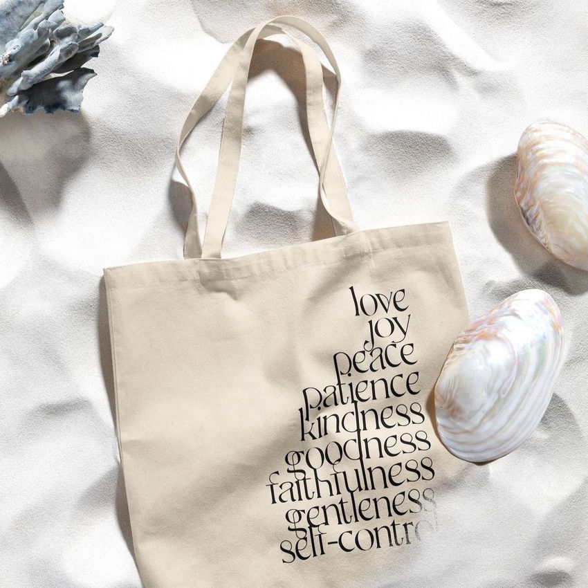 Tote bag mockup featuring a beige fabric bag with black text, placed on light sand with large seashells as props.