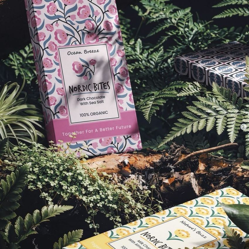 Chocolate box mockup featuring floral packaging designs placed among green ferns and forest foliage in natural light.