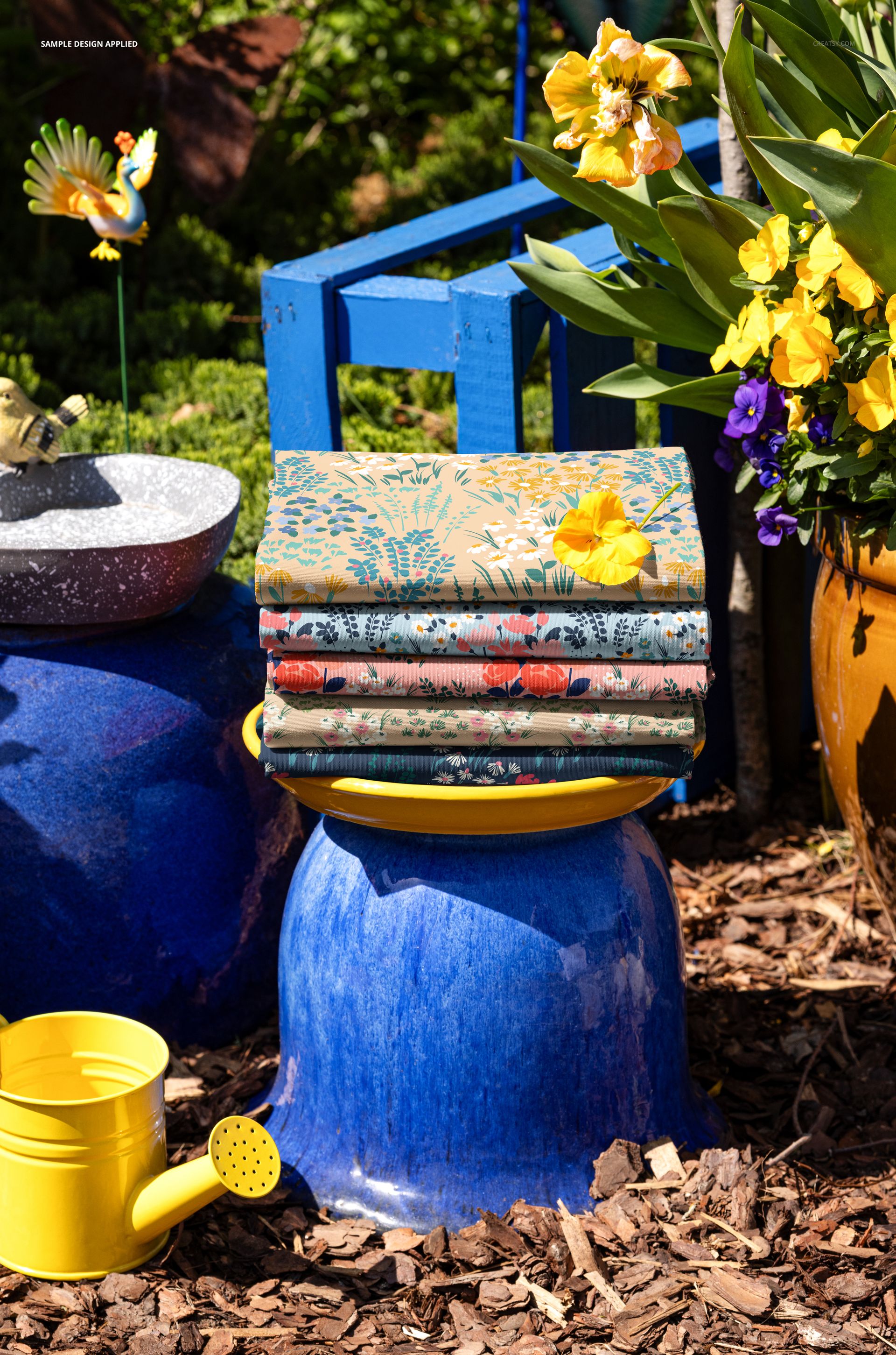 Patterned textile mockup featuring stacked fabrics on a ceramic pot, with bright flowers and garden accessories nearby.