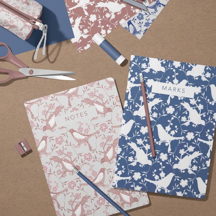 Stationery mockup featuring two classic A4 notebooks with bird and floral patterns, surrounded by pencils, scissors, and other desk items on a brown surface.