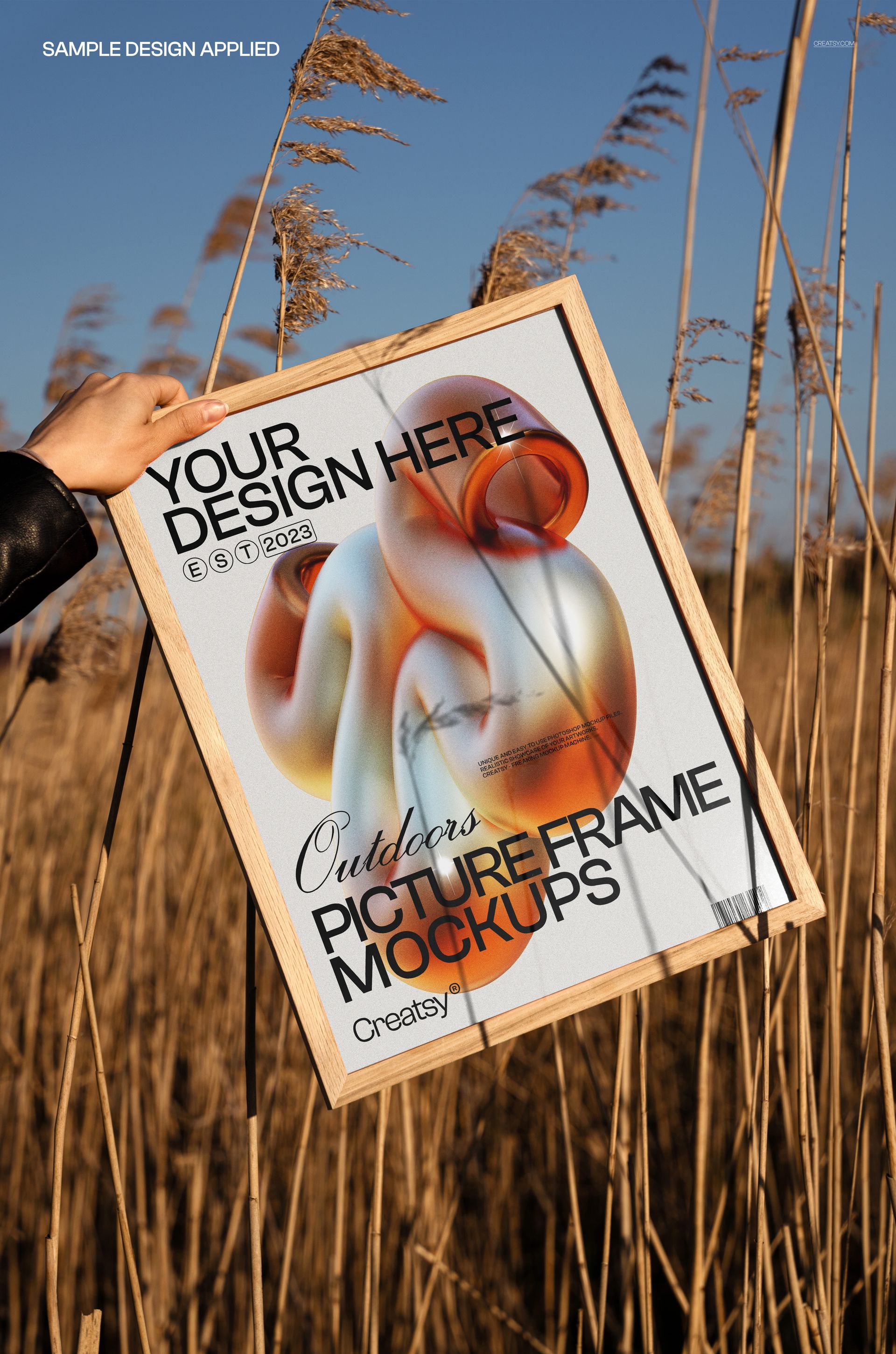 Hand holding a wooden picture frame mockup with abstract art print in tall golden grass under a clear blue sky.