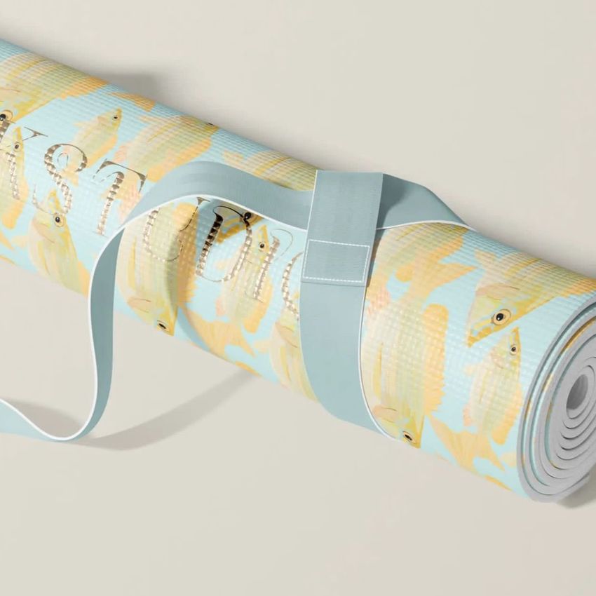 Rolled yoga mat mockup with a light blue strap, featuring a repeating yellow fish pattern on a pale background.