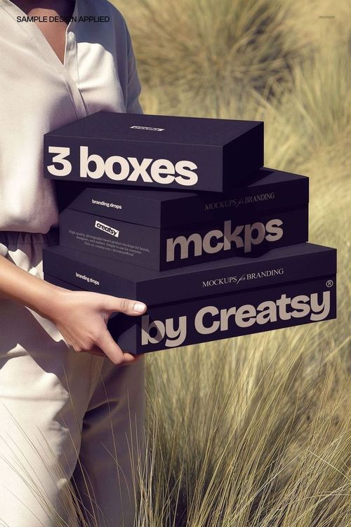 Person holding three stacked rectangular packaging boxes with bold text, standing outdoors among tall grass.