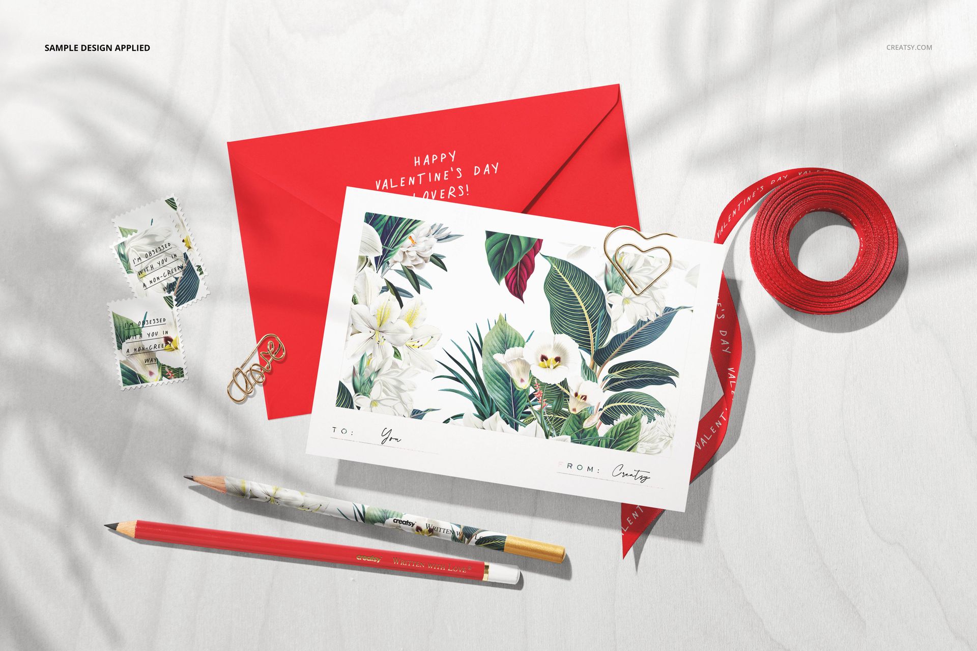 Valentine’s stationery mockup featuring a red envelope, floral card, pencils, ribbon, stamps, and clips on a light surface.