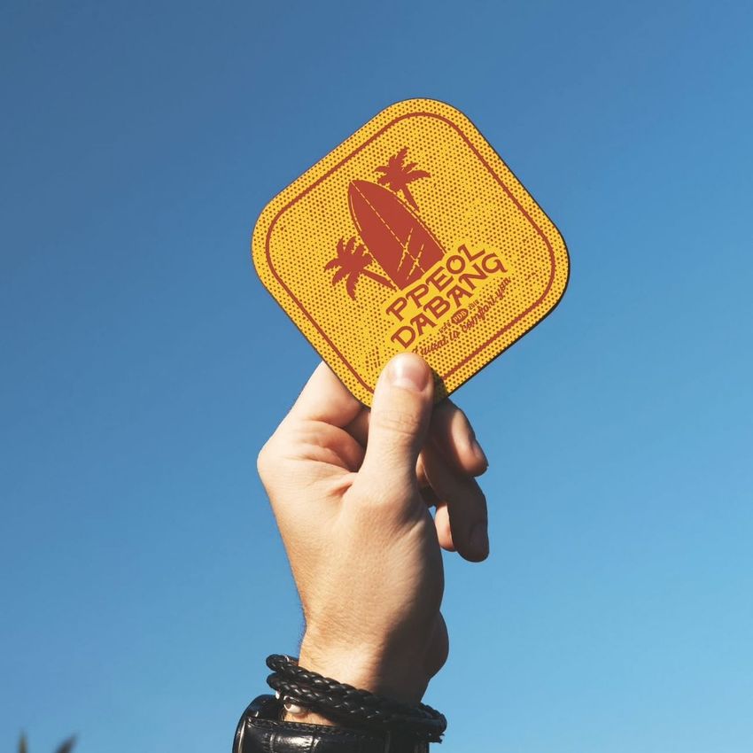 Hand holding a yellow square coaster mockup with red graphic and text against a clear blue sky background.
