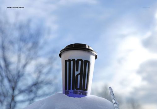Winter-themed PSD mockup of a paper cup with a dark lid, placed on snow against a sky and tree backdrop.