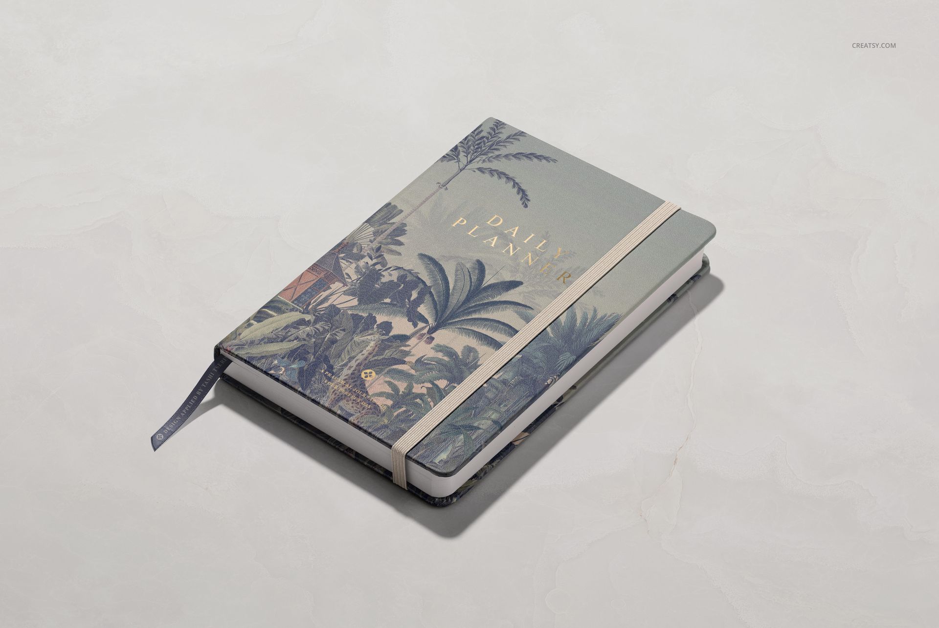 Front angled view of a leather notebook mockup featuring tropical leaf design and elastic closure on a pale background.