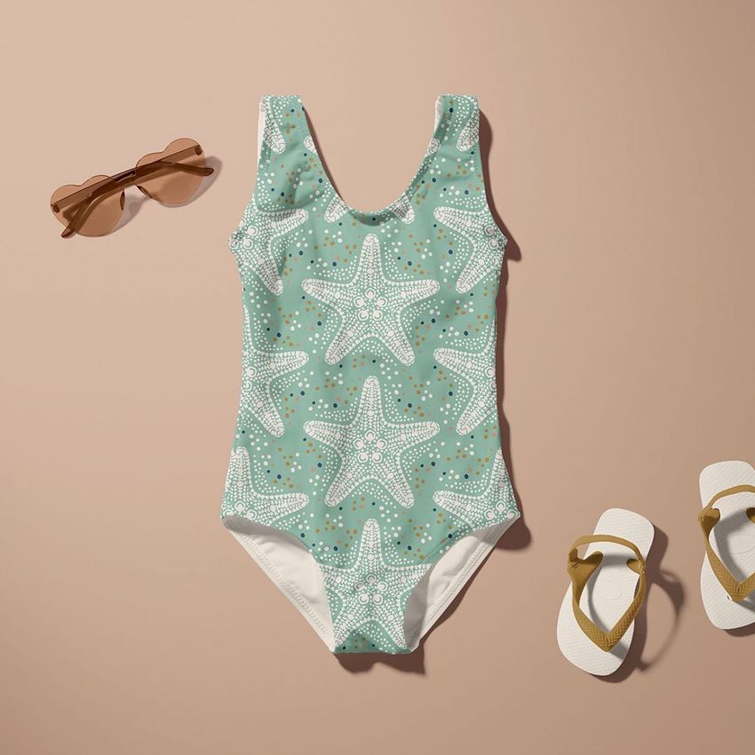 Baby swimsuit mockup featuring a seafoam green one-piece with white starfish pattern, placed on a beige surface with sunglasses and flip-flops.