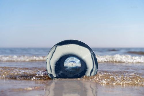 Blue and white patterned vinyl record mockup displayed on the shoreline, partially submerged in shallow water.