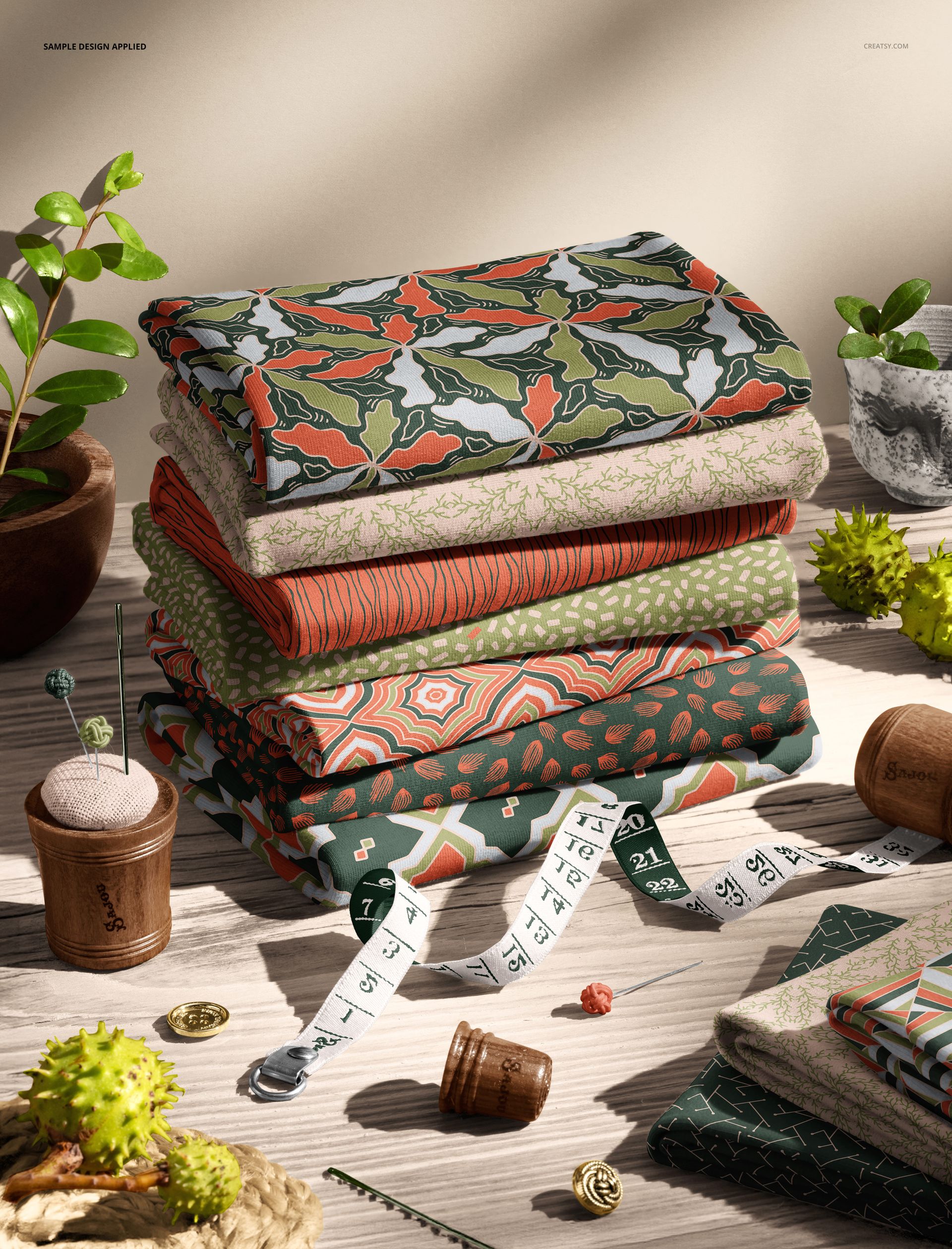 PSD mockup showing folded jersey fabrics with various designs, surrounded by greenery and sewing accessories.