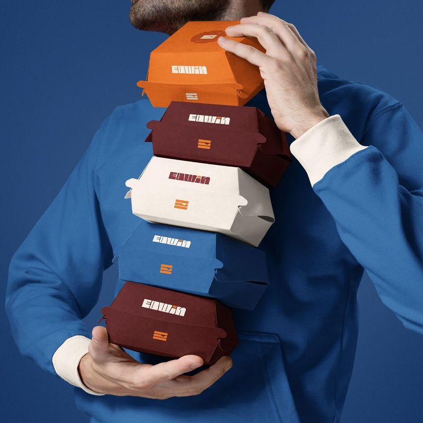 Person in blue hoodie holding a stack of colorful burger boxes mockup against a solid blue background, with visible text overlays.