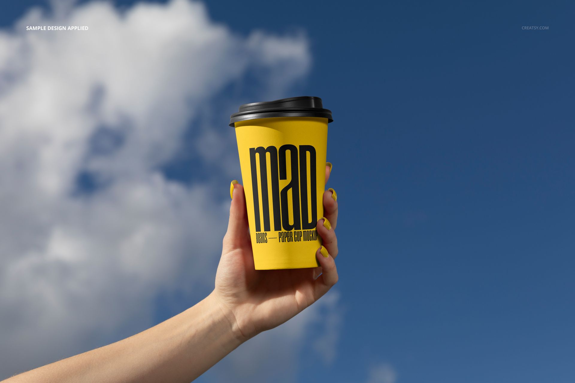 Front view of a yellow paper cup mockup with black lid and large text, held by a hand in an outdoor urban setting.