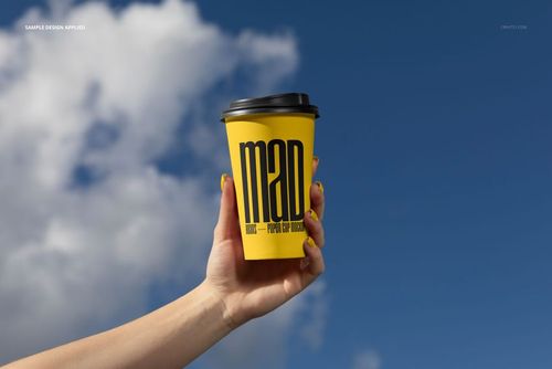 Front view of a yellow paper cup mockup with black lid and large text, held by a hand in an outdoor urban setting.