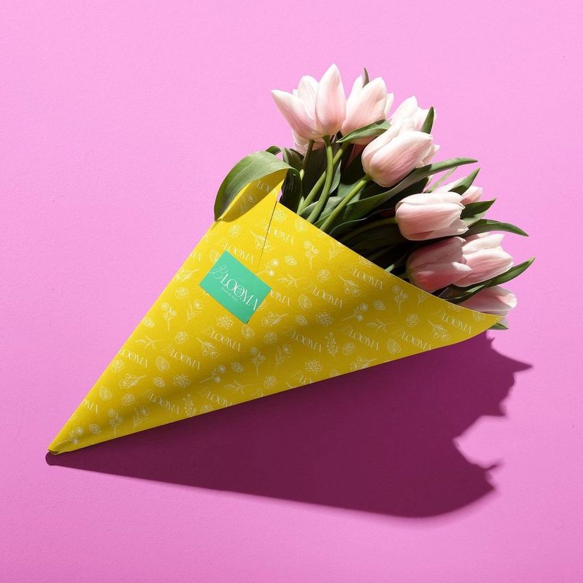 Bouquet of light pink tulips wrapped in yellow patterned packaging paper with a green tag, placed on a bright pink background.