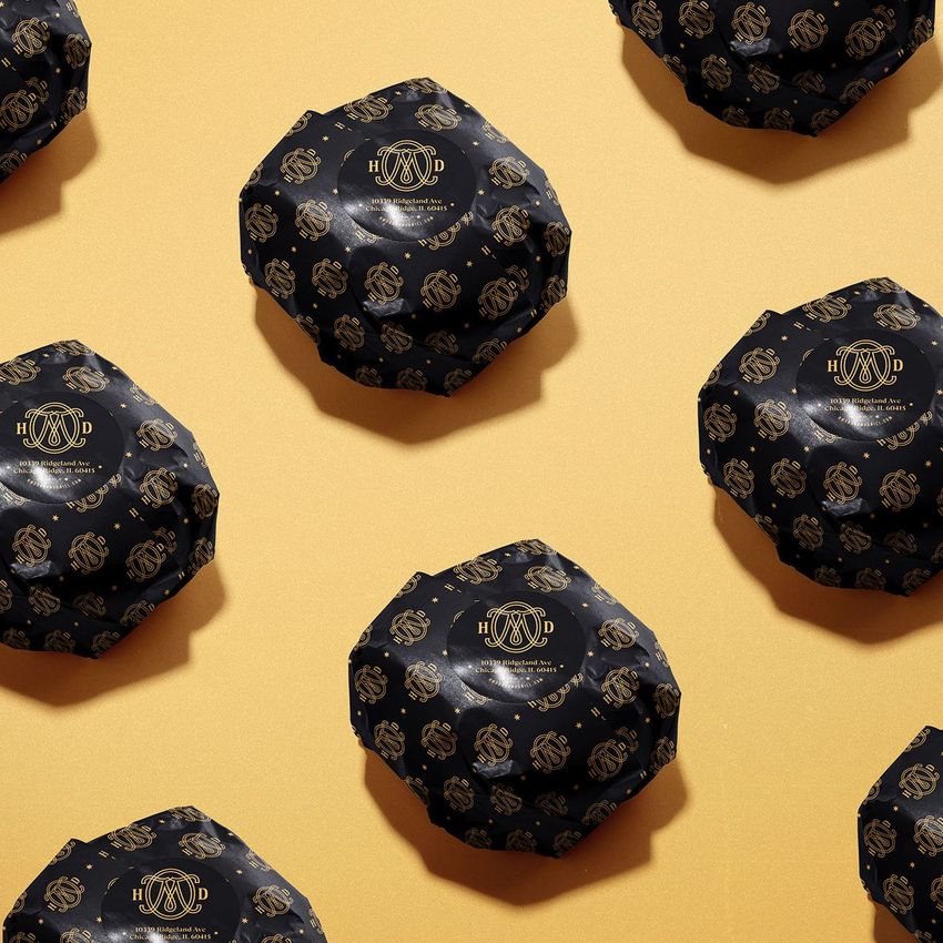 Burger mockup featuring multiple round burgers wrapped in dark patterned paper with gold logos, arranged on a warm yellow surface.
