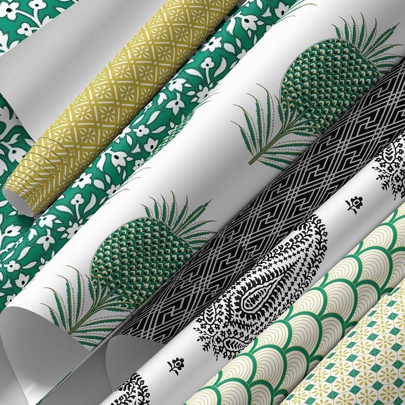 Paper wallpaper mockup showing several rolled sheets with geometric, floral, and pineapple patterns in green, gold, and black.