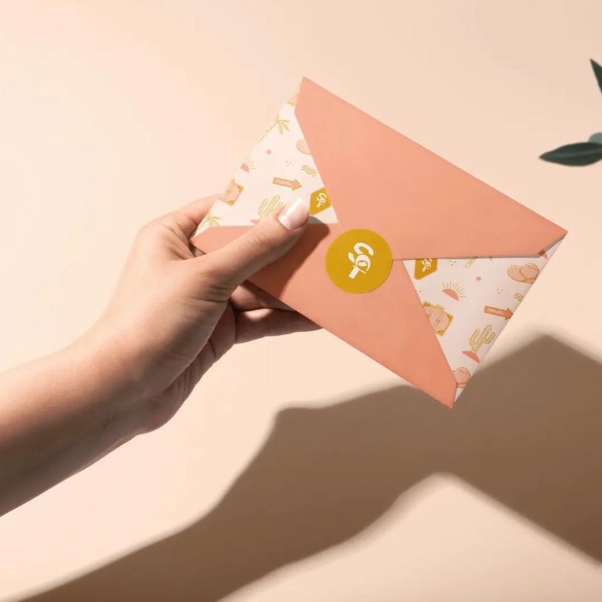 Close-up of a hand presenting a stationery envelope mockup with a colorful design and sticker seal, soft shadows on beige surface.