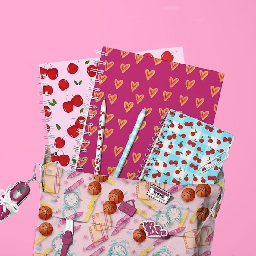 School backpack mockup with spiral notebooks featuring colorful cherry, heart, and geometric patterns on a pink background.