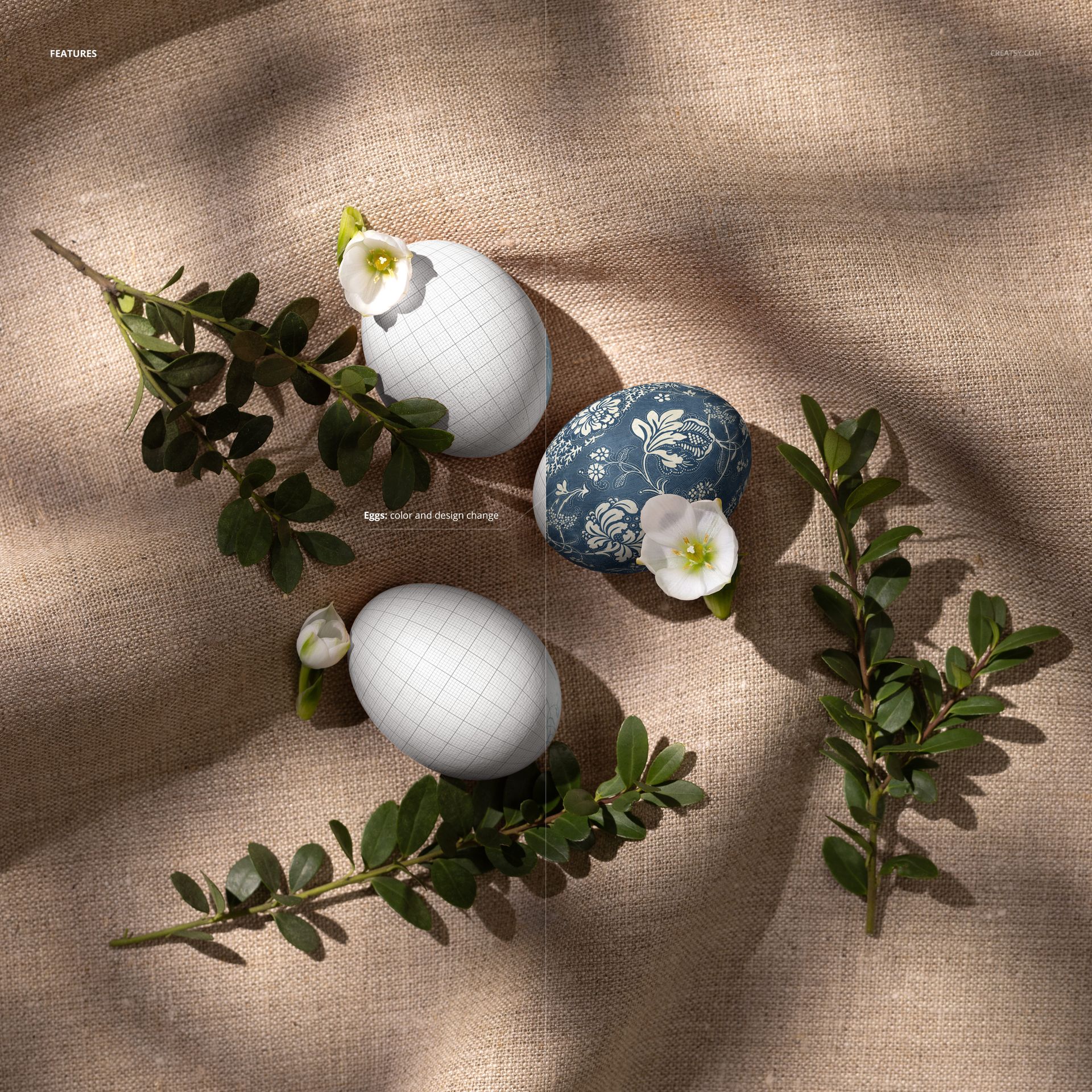 Three Easter eggs arranged on natural linen fabric with white flowers and boxwood branches, top view mockup.