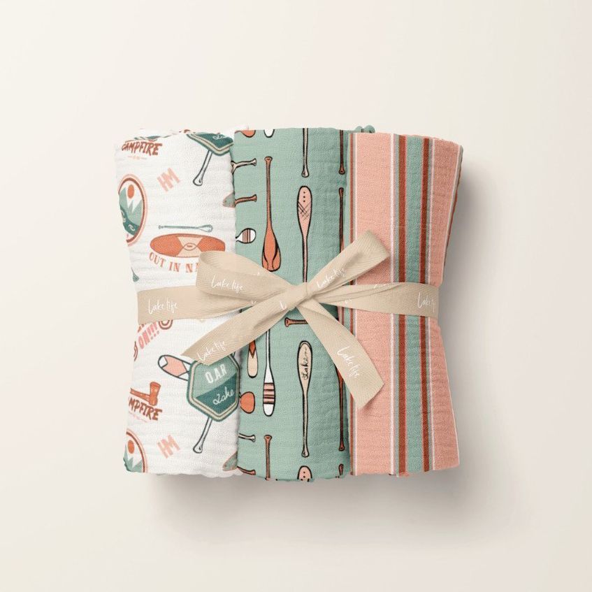 Three crinkle muslin swaddle blankets are folded and tied with a beige ribbon, featuring playful baseball and striped patterns.