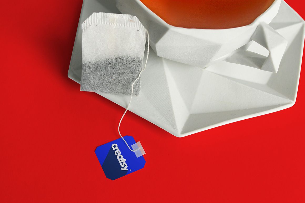 PSD mockup featuring a tea bag and label beside a white cup and angular saucer, all placed on a red background.