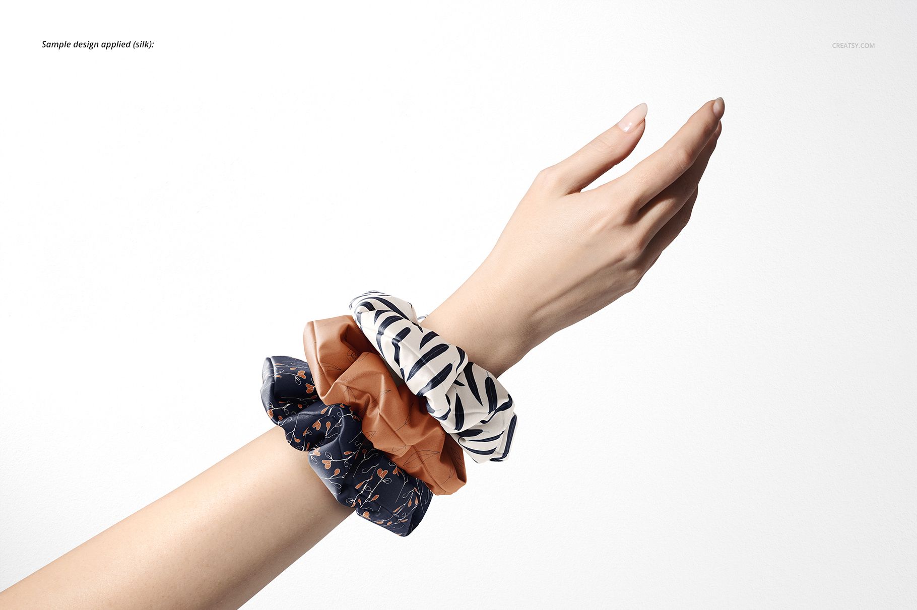 Fashion accessories mockup featuring a hand wearing three hair scrunchies in different colors and patterns on a plain background.