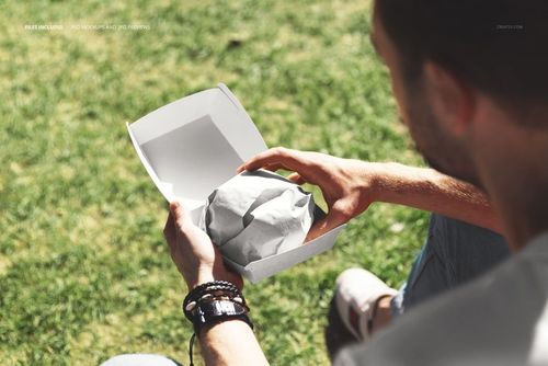 Outdoor scene showing a white packaging box with a wrapped burger being held over green grass, PSD mockup style.