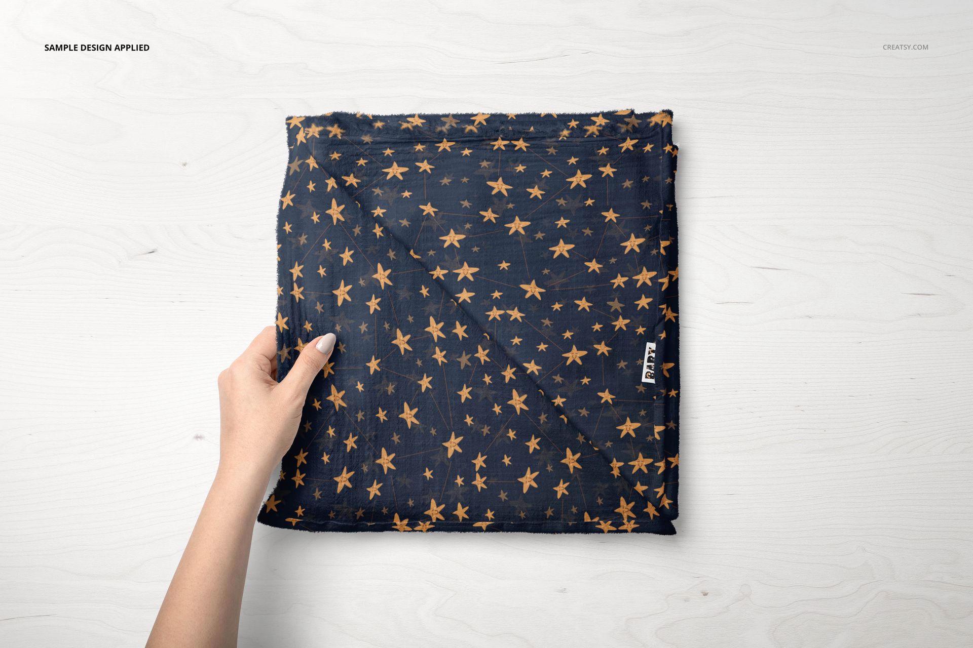 Top view of a muslin swaddle blanket mockup with scattered orange star design, held by hand on light wood background.