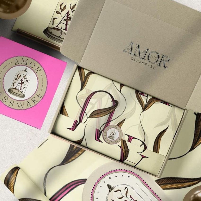 Mailing box mockup featuring an open cardboard box with 'AMOR GLASSWARE' text, decorative wrapping paper, and a round sticker.
