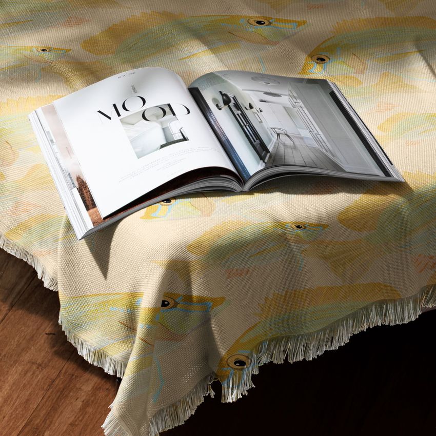 Polyester woven coverlet mockup draped over a wooden surface with a fringed edge, featuring a fish pattern and an open magazine.
