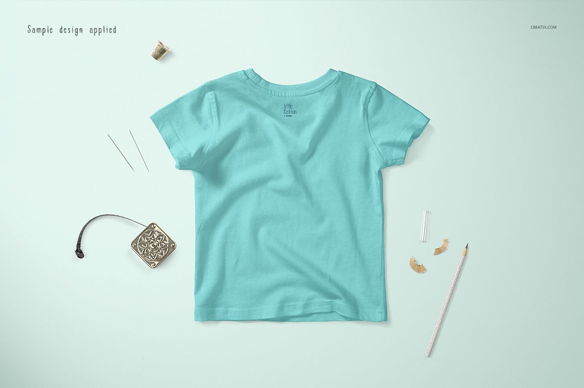 Light blue baby T-shirt mockup shown from the back, placed on a soft blue background with sewing accessories nearby.