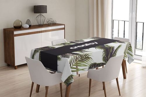 Dining table covered with a mockup tablecloth featuring green palm leaves and a solid black runner in a bright interior.