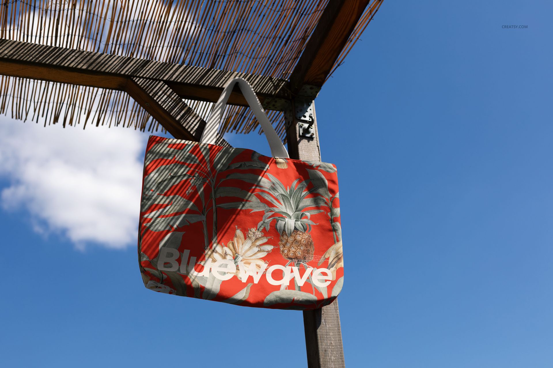Tote bag mockup featuring a red and green tropical design with pineapples, displayed against a clear blue sky.