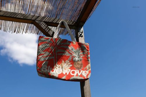Tote bag mockup featuring a red and green tropical design with pineapples, displayed against a clear blue sky.