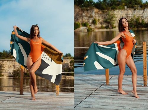 Woman standing on a wooden deck by a lake holding a patterned beach waffle towel, wearing an orange one-piece swimsuit.