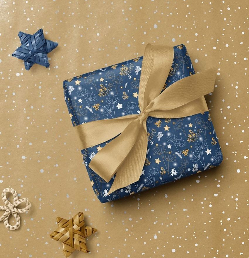 Square gift box mockup wrapped in blue paper with gold and white festive patterns, tied with a gold ribbon, on a brown speckled background.